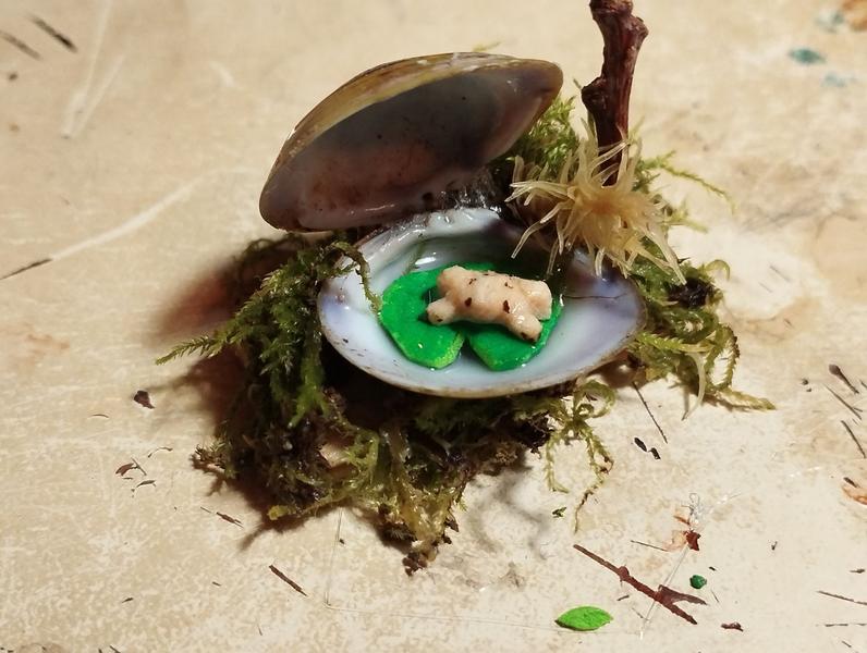 Une mini grenouille en bois sur un nénuphar. Le nénuphar est dans un coquillage bivalve entouré de mousse