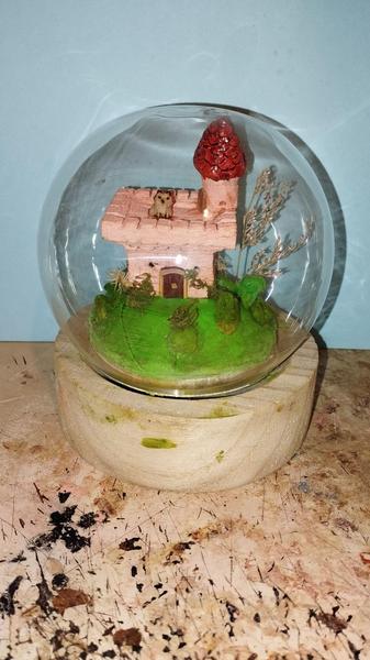 Une scène miniature avec un château dans une boule en verre. Le château à un simple donjon et une tourelle. Il surplombe une colline verdoyante. Un renard veille en haut du donjon