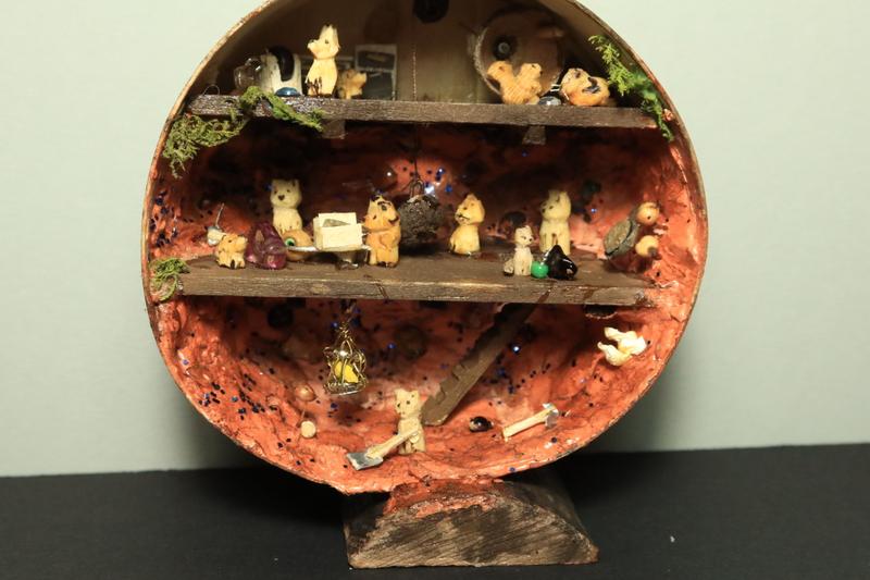 Photo d'une boîte ronde transformée en mine. Il y a trois étages. Au sous sol, un chat creuse avec une pelle, un canari est enfermé dans une cage.Au milieu, plusieurs chats et renards travaillentÀ l'étage du haut, un cochon d'inde et un écureuil font tourner une roue, des renards meulent des pierres 