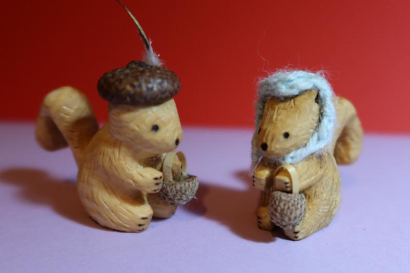 Deux sculptures d'écureuil en bois. Les deux tiennent un panier fabriqué avec une cupule de gland.Celle de gauche porte un chapeau cupule avec une plume dorée et argentée. Celui de droite porte un capuchon crocheté bleu.