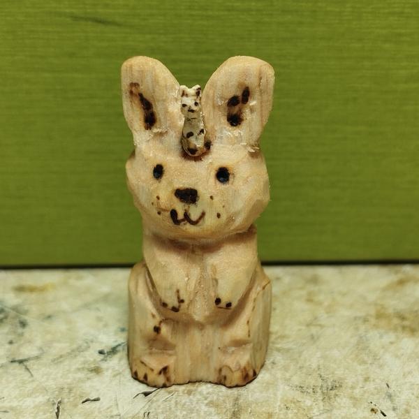Une sculpture de lapin en bois debout sur ses pattes arrières. Un mini lapin est logé entre ses oreilles. 