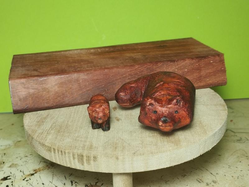 Deux pandas roux en bois. Un petit et un plus grand. Le plus grand est allongé sur ses pattes. Le bois est de couleur rouge foncé.Derrière, un morceau de planche de padouk