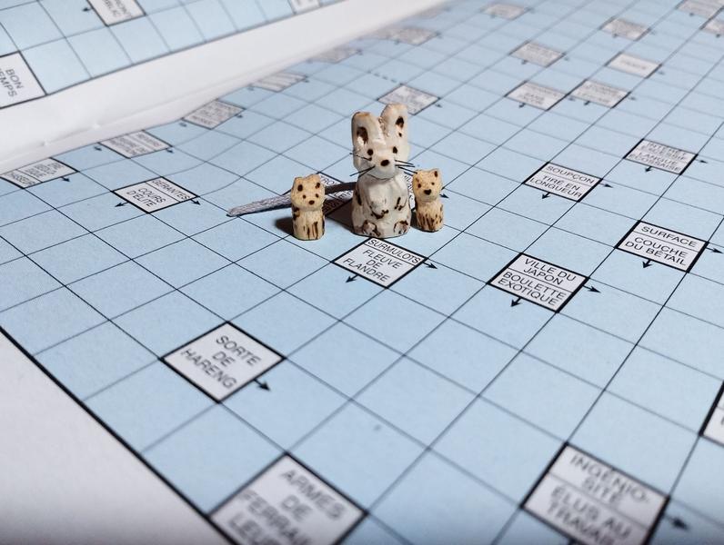 Mini sculptures en bois : deux chats et une souris, un peu plus grande. Elles sont posées sur une grille de mots fléchés à côté de la définition "surmulots" (4 lettres) 