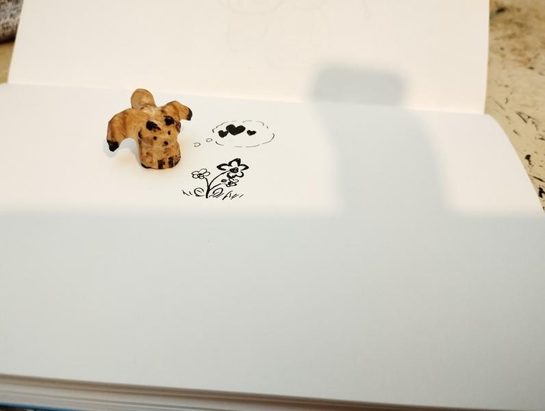 A sculpture of a kitsune, a fox with 3 tails, looking at a drawing of 3 flowers sharing the same base. A cartoon balloon is showing 3 hearts next to the fox 