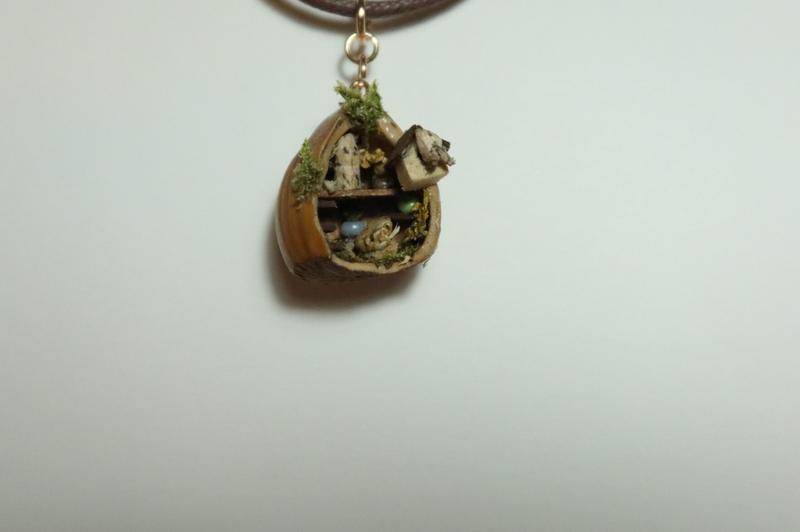 Un pendentif fait dans une noisetteIl représente un petit coin de verdure, avec un petit chat, un nichoir, un oiseau et une chaise