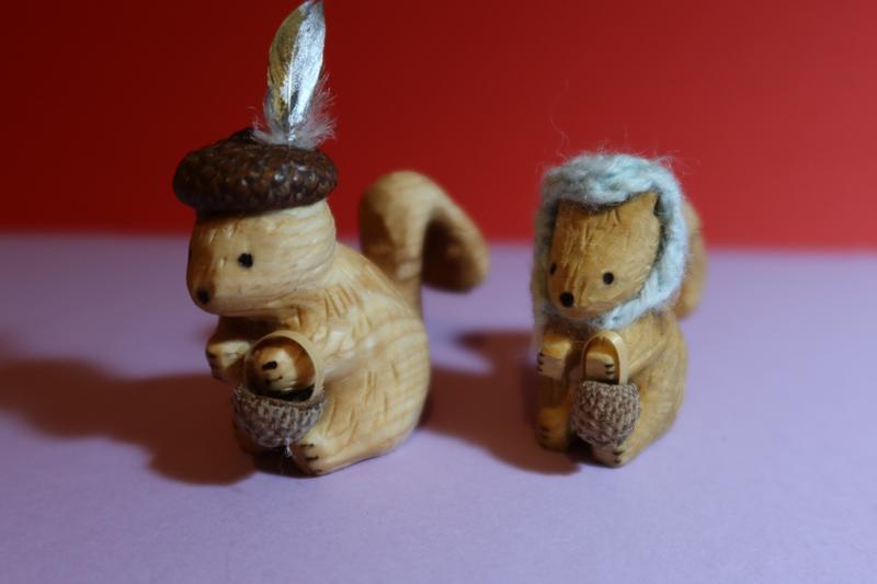 Deux sculptures d'écureuil en bois. Les deux tiennent un panier fabriqué avec une cupule de gland.Celle de gauche porte un chapeau cupule avec une plume dorée et argentée. Celui de droite porte un capuchon crocheté bleu.