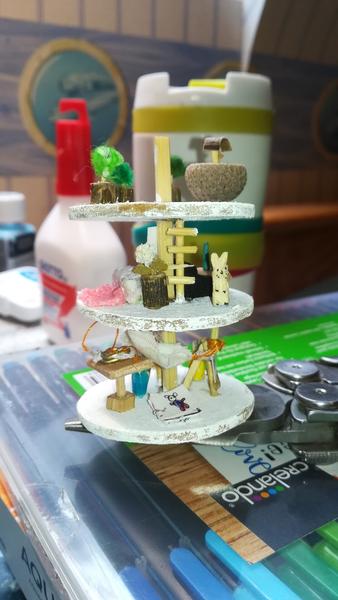 Vue de l'atelier hors du pot. Il y a trois étages relié par une tige centrale. On distingue un mini lapin, une échelle, des pots de fleurs, et des mini tableaux