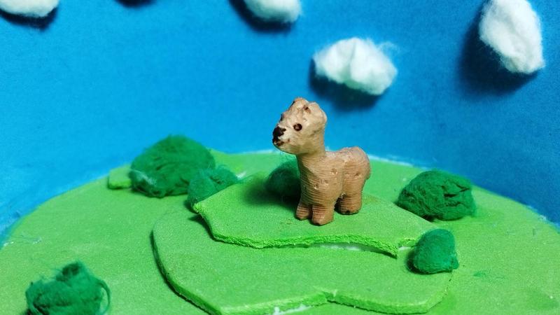 Photo d'une scène miniatureUn petit alpaga en bois marche sur une prairie verte parsemée de quelques buissons. Il sourit. En fond un ciel bleu où s'élèvent quelques nuages