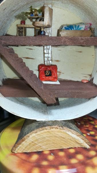 Une mini maison en bois en forme de rond. Le diamètre est de 8cm. Au rez-de-chaussée il y a une cheminée avec un mini calcifer. Au premier il y a un lit, un bureau, un fauteuil, des plantes et un mini renard. 