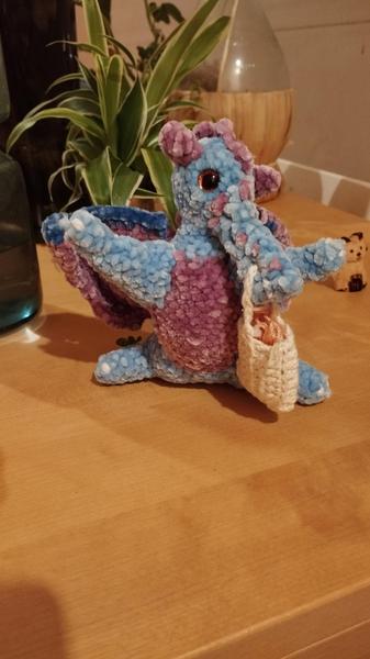 Un dragon bleu et violet au crochet. Il tient un sac dans sa gueule