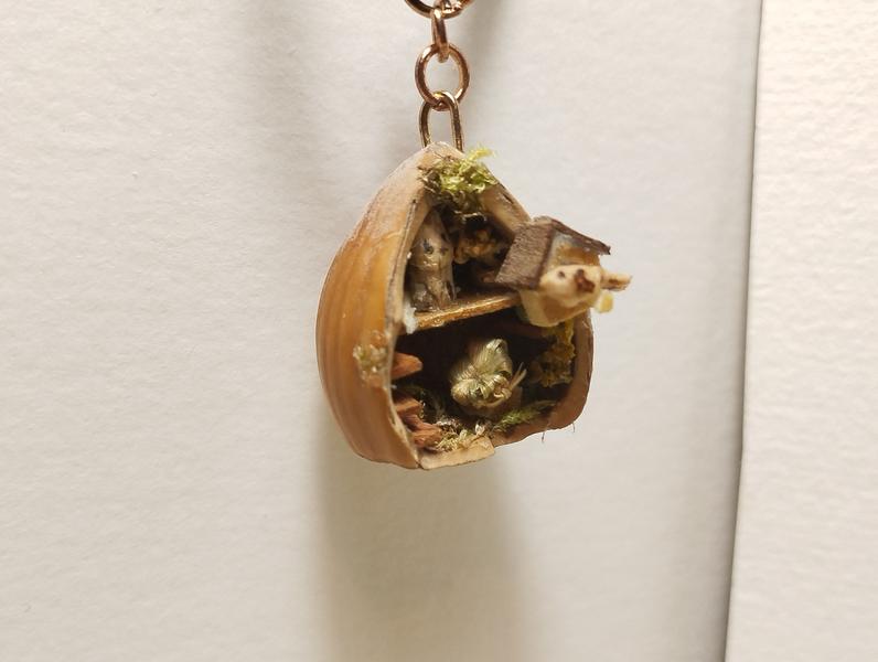 Un pendentif noisette. Dans le creux de la noisette il y a une mini sculpture de chat, un fauteuil et de la mousse. Sur l'un des rebords, il y a un nichoir avec un petit oiseau