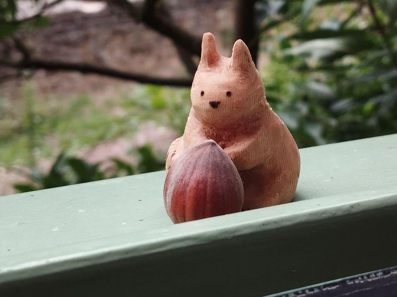 Un écureuil en bois qui tient une noisette. L'écureuil est vu de face, il a de grandes oreilles. L'écureuil est deux fois plus grand que la noisette