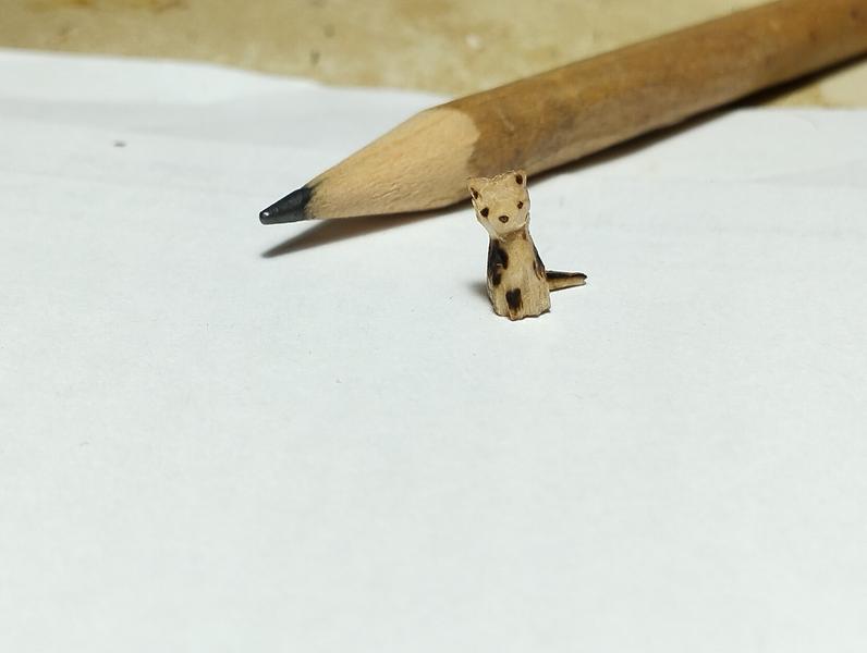 Mini sculpture de chat en bois, à côté d'un crayon gris posé à plat . Le chat, assis, est un peu plus haut que le crayon