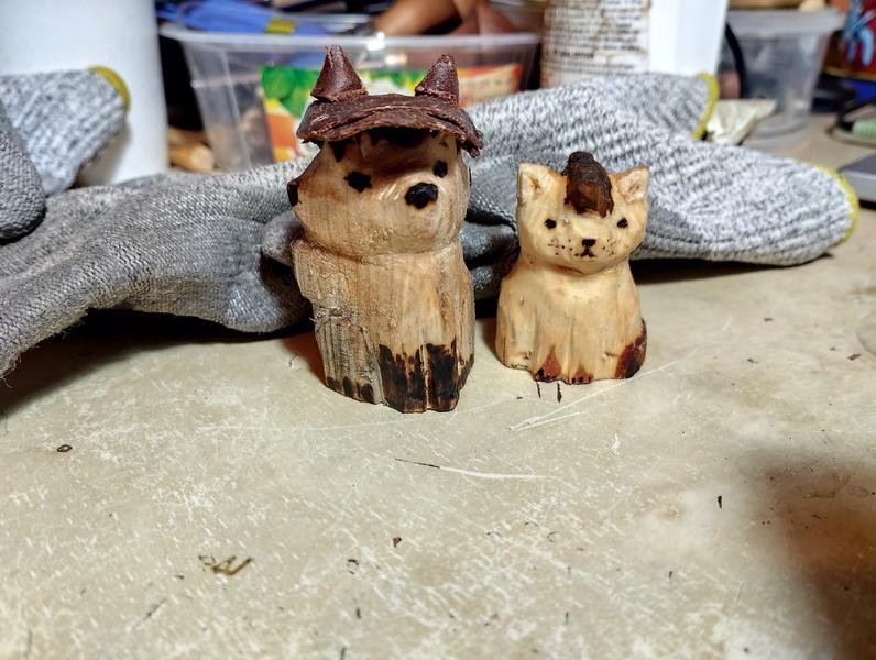 Deux sculptures en bois : un renard et un chat. Le renard porte un béret en cuir. Le béret a deux pointes pour les oreilles. Le chat porte une crête. 