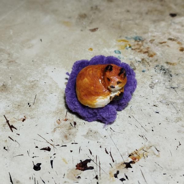 Un petit renard en bois qui dort sur un coussin violet. Le renard est teint en orange