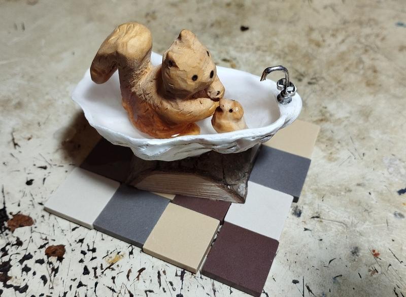 Photo d'un petit écureuil en bois dans une baignoire. Il joue avec un canard. La baignoire est une coquille d'huître 