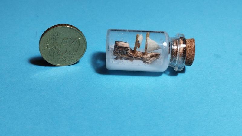 Photo d'un petit bateau en bois dans une fiole. À côté une pièce de 50 centimes, son diamètre est un peu plus grand que celui de la fiole