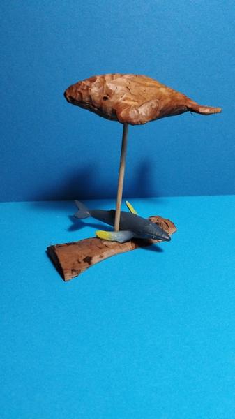 Une sculpture de baleine en bois. La baleine est posée sur une tige. Sur le socle, il y a une baleine en plastique 