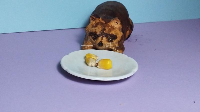 Sculpture en bois d'un raton laveur. Vu de face, il pose sa truffe sur une assiette de maïs
