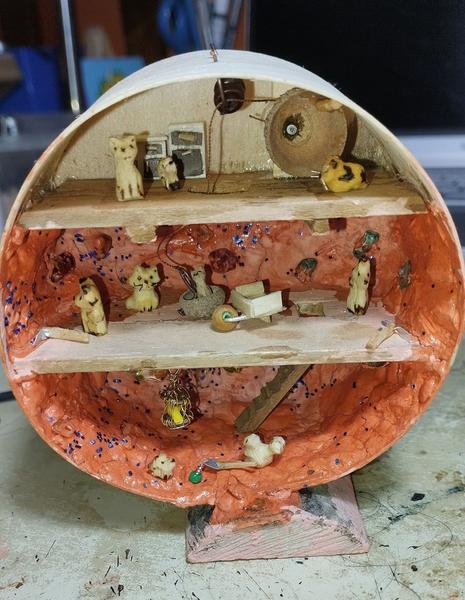 Photo d'une mine dans une boite ronde en bois.Il y a trois étages, celui du haut est aménagé, ceux du bas ont les parois recouvertes de terre orange pailletée.Des petits animaux en bois (chat, renard, écureuil, etc) s'activent dans la mine